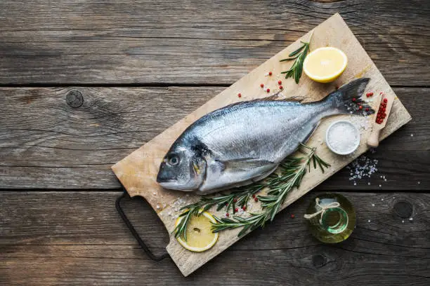 Photo of Fresh dorado fish with lemon slices,  sea salt and rosemary twigs on wooden cutting board. Top view. Copy space for text.