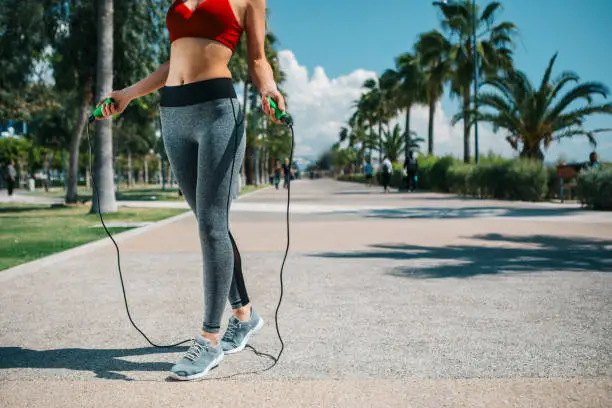 Low angle close up of slim female athlete body using a skipping rope. Woman is standing on alley along palm tree park. Copy space