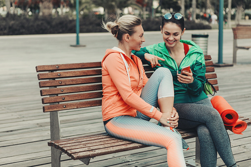 Happy two girls are talking and laughing while sitting on bench outdoor. Lady is showing smartphone to her friend. They are wearing sportswear