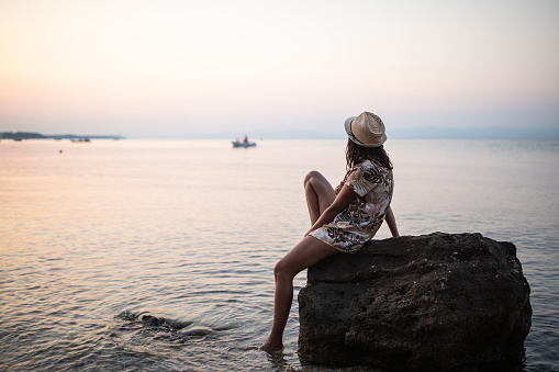 Young  woman enjoying alone by the sea on sunset, sitting on rock with barefoot in the water