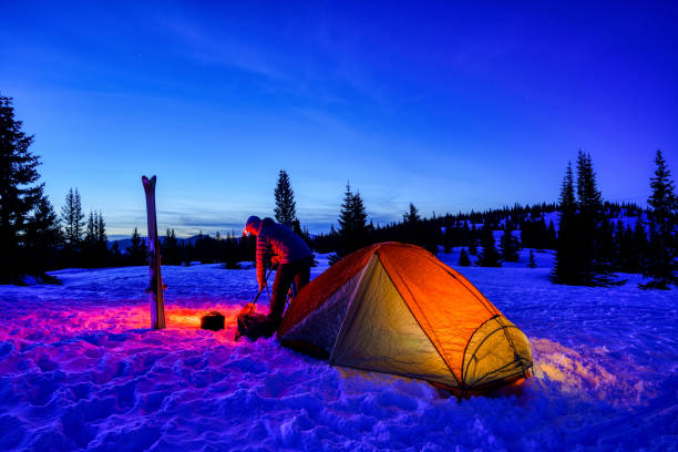 Winter Ski Camping in the Mountains - Glowing orange tent at ski base camp for touring in the mountains with gorgeous mountain backdrop. Man shoveling snow around camp.