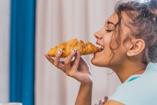 Photo of a cheerful young woman happy. Going to the table and eating croissant. Looking only at the croissant.