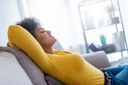 People, rest, comfort and leisure concept - African american young woman resting on sofa at home. Relaxed woman lying on comfortable sofa on pillows with hands behind head in living room.