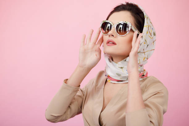 Stylish confident brunette woman touching his sunglasses, wearing in scarves, looking up, isolated on a pink background. stock photo