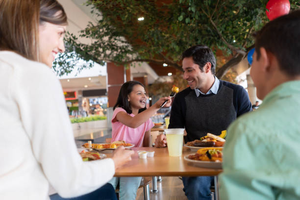 Happy girl eating luch with her family and sharing with her father Happy girl eating luch with her family and sharing food with her father while smiling - lifestyle concepts food court photos stock pictures, royalty-free photos & images