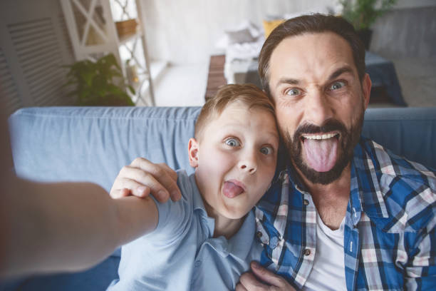 Outgoing parent and child grimacing on camera Portrait of happy boy and optimistic dad shoving tongues while taking selfie grimacing photos stock pictures, royalty-free photos & images