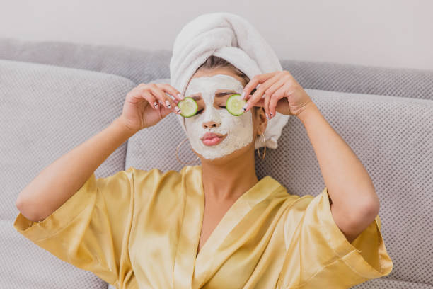A beautiful young woman gets a face mask in the spa center, lying with cucumbers in her eyes. Happy and gets a lot of fun. Beautiful young woman doing spa treatments at home, lying on the couch. Holds pieces of cucumbers in the arms above the eyes. Dressed in a bathrobe and a towel on the head. cucumber stock pictures, royalty-free photos & images