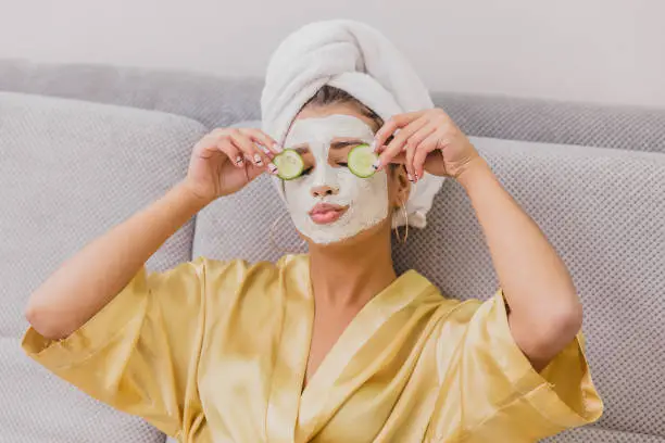 Beautiful young woman doing spa treatments at home, lying on the couch. Holds pieces of cucumbers in the arms above the eyes. Dressed in a bathrobe and a towel on the head.