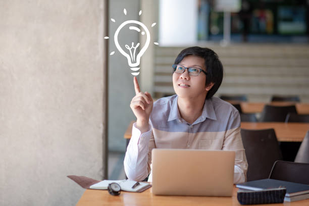 istock Young Asian man university student with glasses pointing at light bulb graphic illustration while using laptop computer for learning in college library. Creative thinking idea and inspiration concepts 1138651367
