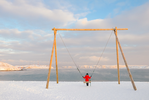 Single young Chinese woman sitting on a huge yellow swing on beach by Arctic ocean, in Teriberka, Russia