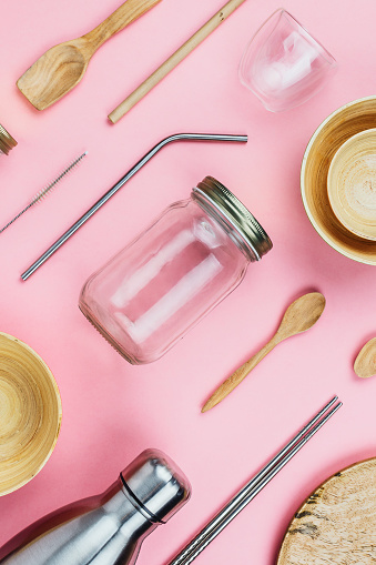 Flatlay of various sustainable zero waste kitchenware utensils and dishware: glass and metal bottles, wooden bamboo bowls, eco straws etc on pink