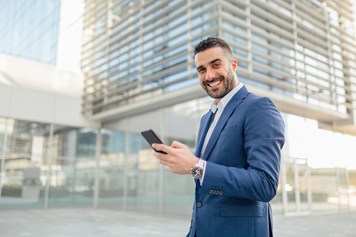 Young businessman posing in front of the building, holding the phone and smiling