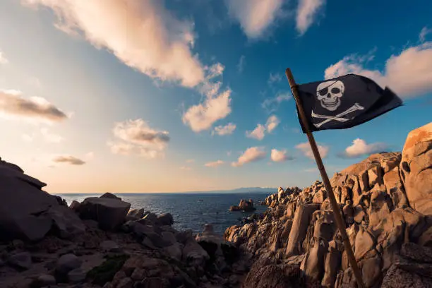 jolly roger flag flies at sunset pinned between the rocks of the pirate island in front of a few clouds in the crystal clear blue and orange sky, Capo Testa, Santa Teresa di Gallura, Sardinia, Italy