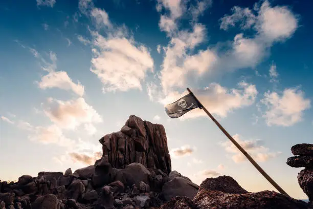 jolly roger flag flies pinned between the rocks of the pirate island in front of a few clouds in the clear blue sky, Capo Testa, Santa Teresa di Gallura, Olbia Tempio, Sardinia, Italy