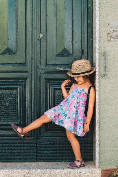 Smiling little girl lifts a leg next to the green door of her house, she wears a lilac summer dress of flowers and a straw hat, vertical image