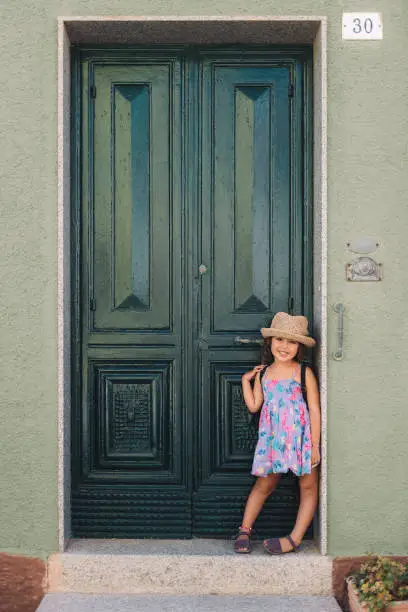 Little girl poses smiling leaning against the green door of her house, she wears a lilac summer dress of flowers and a straw hat, vertical image