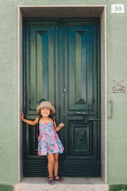 Little girl poses funny and smiling in front of the green door of the house, she wears a lilac summer dress of flowers and a straw hat, vertical image