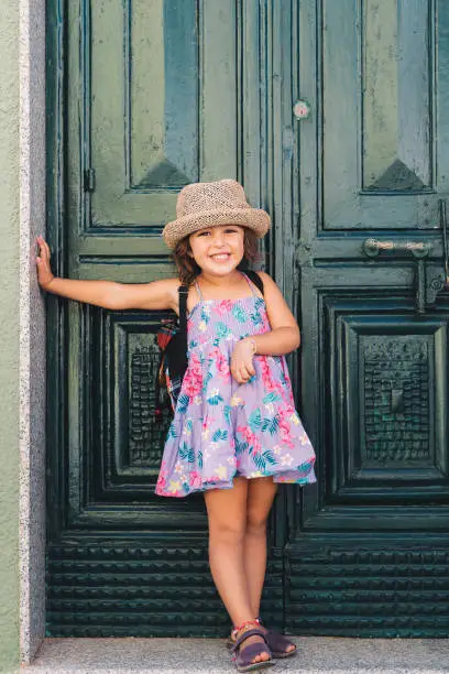 Little girl poses amused in front of the green door of house, wears a lilac summer dress of flowers and a straw hat, vertical image