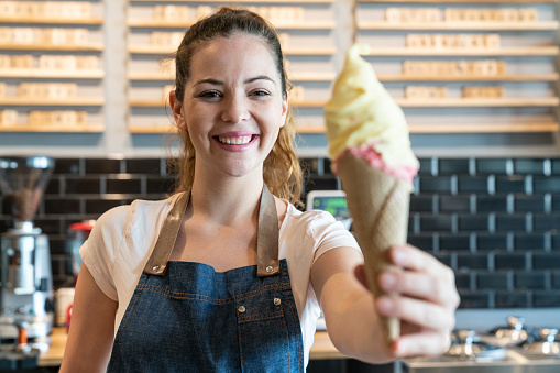 Portrait of friendly woman working at an ice cream parlor handing an ice cream while smiling at camera very happy