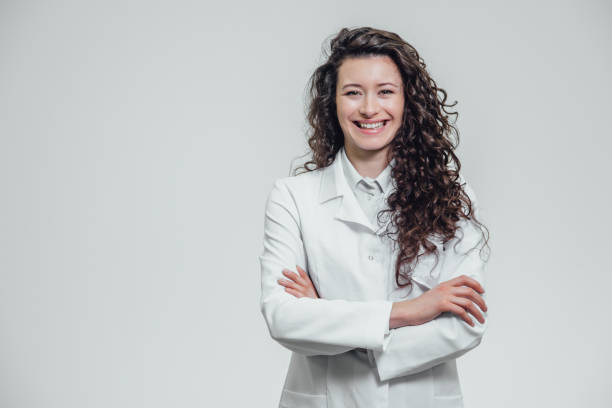 ritratto di felice giovane sorridente ragazza dottore. vestito con una veste bianca. uniformemente in piedi con le mani incrociate su uno sfondo grigio. - camice da medico foto e immagini stock