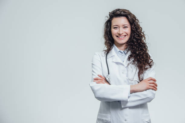 ritratto di felice giovane sorridente ragazza dottore. vestito con una veste bianca. uniformemente in piedi con le mani incrociate su uno sfondo grigio. - medicine lab coat hospital senior adult foto e immagini stock