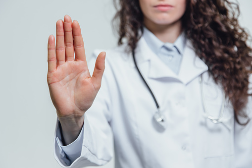 A young beautiful woman doctor gesturing stops. Probably a female doctor shows a gesture stop over a gray background. Has beautiful long black curly hair.