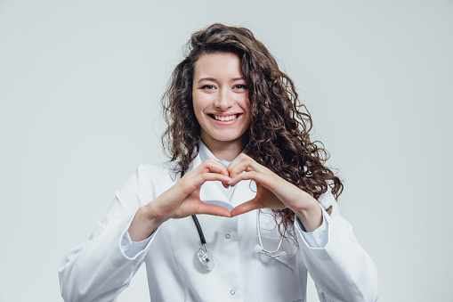 A young woman doctor is on a gray background. During this time, the heart symbol shows on the chest with his hands. Dressed in a white robe. Female doctor with a stethoscope gesturing. Has beautiful long black curly hair.