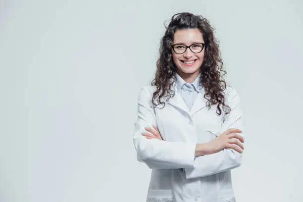 Photo of Portrait of smiling young female doctor. Beautiful brunette in white medical gown in glasses. Holding without a stethoscope on a gray background.