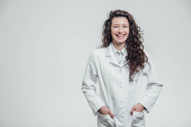 retrato horizontal da menina de um doutor bonito novo. sorrindo, olhando diretamente para a câmera. em um fundo cinzento, prendendo as mãos em seu bolso e apontando a uma cópia do espaço - jaleco de laboratório - fotografias e filmes do acervo