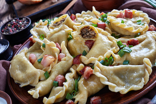hot pierogies or dumpling with ground meat and liver filling sprinkled with spring onion and bacon. sour cream and cutting board with ingredients at the background, view from above, close-up