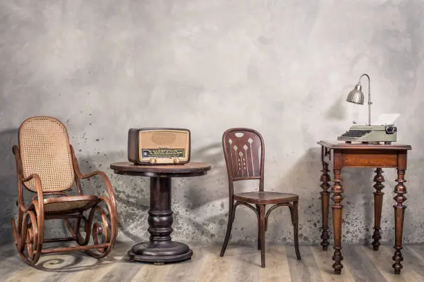 Photo of Vintage loft room with antique rocking chair, broadcast radio, old typewriter and lamp on oak wooden desk front concrete wall background with shadows. Retro style filtered photo