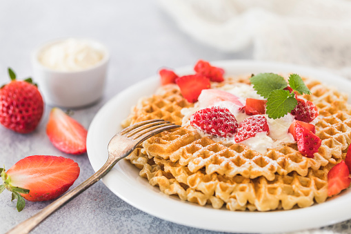 Fresh golden waffles with chopped strawberries and whipped cream on top.  White plate, gray background and a piece of white  fabric next to the plate. There are whole and halfed strawberries next to the plate.