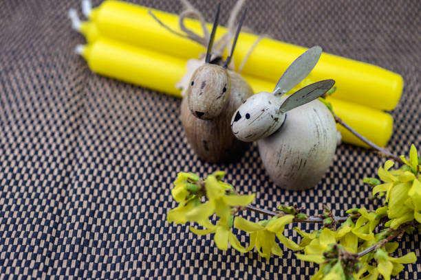 Easter composition on a dark straw mat, with yellow candles and forsythia flowers. The candles are tied with a natural string, and a small wooden bunny is hanging from it