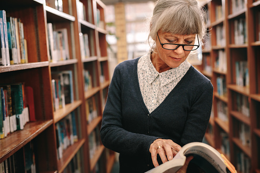 Portrait of an elderly woman reading a book standing in a library beside the book racks. Senior woman going through a reference book in a university library.