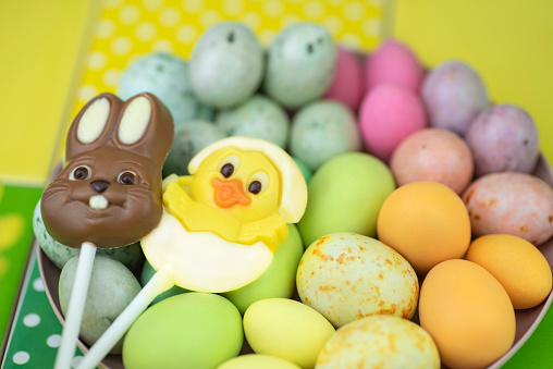Easter composition with two chocolate lollipops over a round pink plate with colorful marzipan Easter eggs.\n\nOne lollipop is formed as a bunny and the other as a chick coming out of its egg.