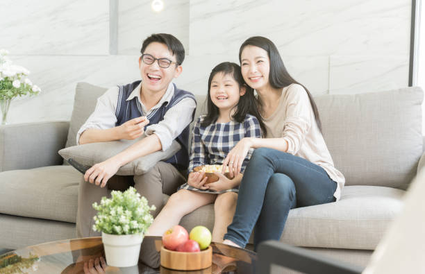 família asiática feliz que presta atenção à tevê junto no sofá na sala de visitas. conceito da família e do repouso. - japanese ethnicity family smiling happiness - fotografias e filmes do acervo