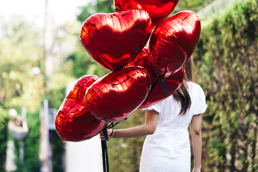 Back view of slim female in white dress carrying bunch of heart shaped balloons while walking on blurred background of park