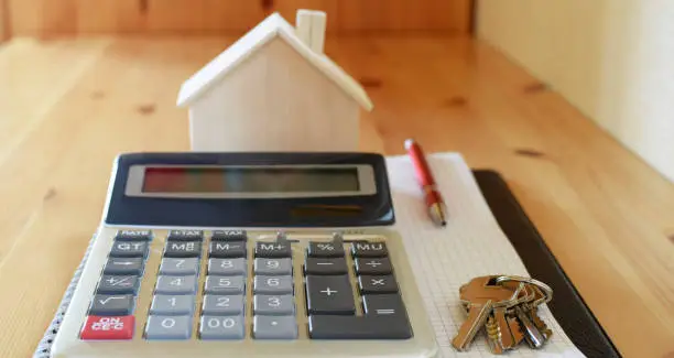 Paper notebook with calculator, house model keys and pen on wooden table.  House building, buying or renting new home planning concept.