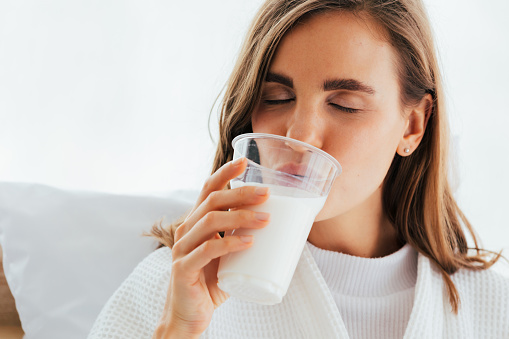 Closeup young attractive woman with closed eyes drinking milk from cup near pillow on white background