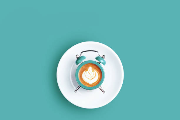 Alarm clock with the dial of cappuccino on turquoise background. Minimal styled coffee time concept.