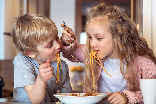 Cute children having fun while eating spaghetti with bolognese sauce at home