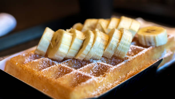Fresh square Belgian waffle with cut banana topping and sugar powder for breakfast on dark background.