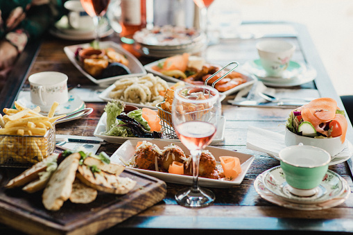 A table filled with delicious looking food served in different sized dishes alongside quirky teacups and glasses of wine.