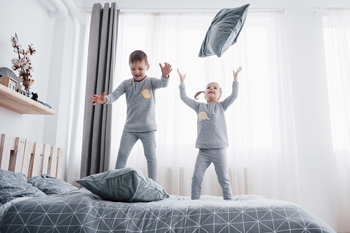 Happy kids playing in white bedroom. Little boy and girl, brother and sister play on the bed wearing pajamas. Nursery interior for children. Nightwear and bedding for baby and toddler. Family at home