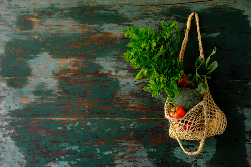 Many colorful contrast color salad vegetables in a reusable string hemp bag hanging from an old weathered abstract color contrasting turquoise colored background wall, with good copy space to the left of the image.