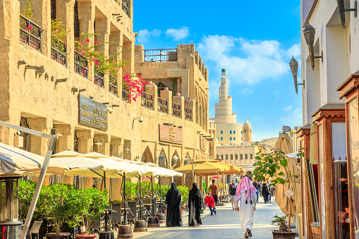 Doha, Qatar - February 20, 2019: women and men in traditional Arab clothing walk along main street inside Souq Waqif old market with cafes and restaurants.Fanar Islamic Cultural Center in the distance