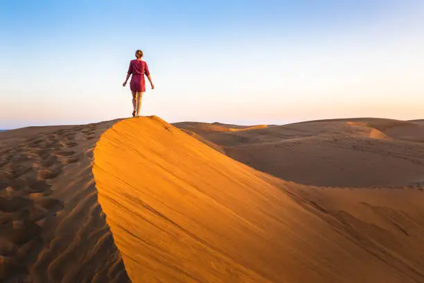 Girl walking on sand dunes in arid desert at sunset and wearing dress, scenic landscape of Sahara or Middle East