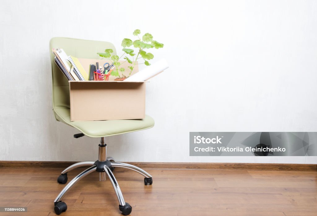 Box with personal items standing on the chair in the office.Concept of moving or dismissal Chair and box with different stuff in it, male hand holding blank speech bubble for your text.Concept of moving/fired from job Employee Stock Photo