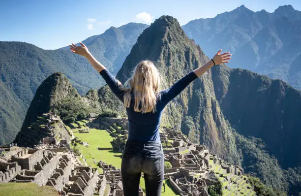 A back view of a blonde young woman, hands in the air, happy to be at Machu Picchu.