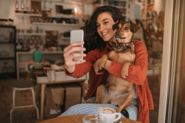Young Woman Sitting In Cafe With Her Dog Young woman sitting in cafe or bookstore with her cute dog and making selfie. photo messaging stock pictures, royalty-free photos & images